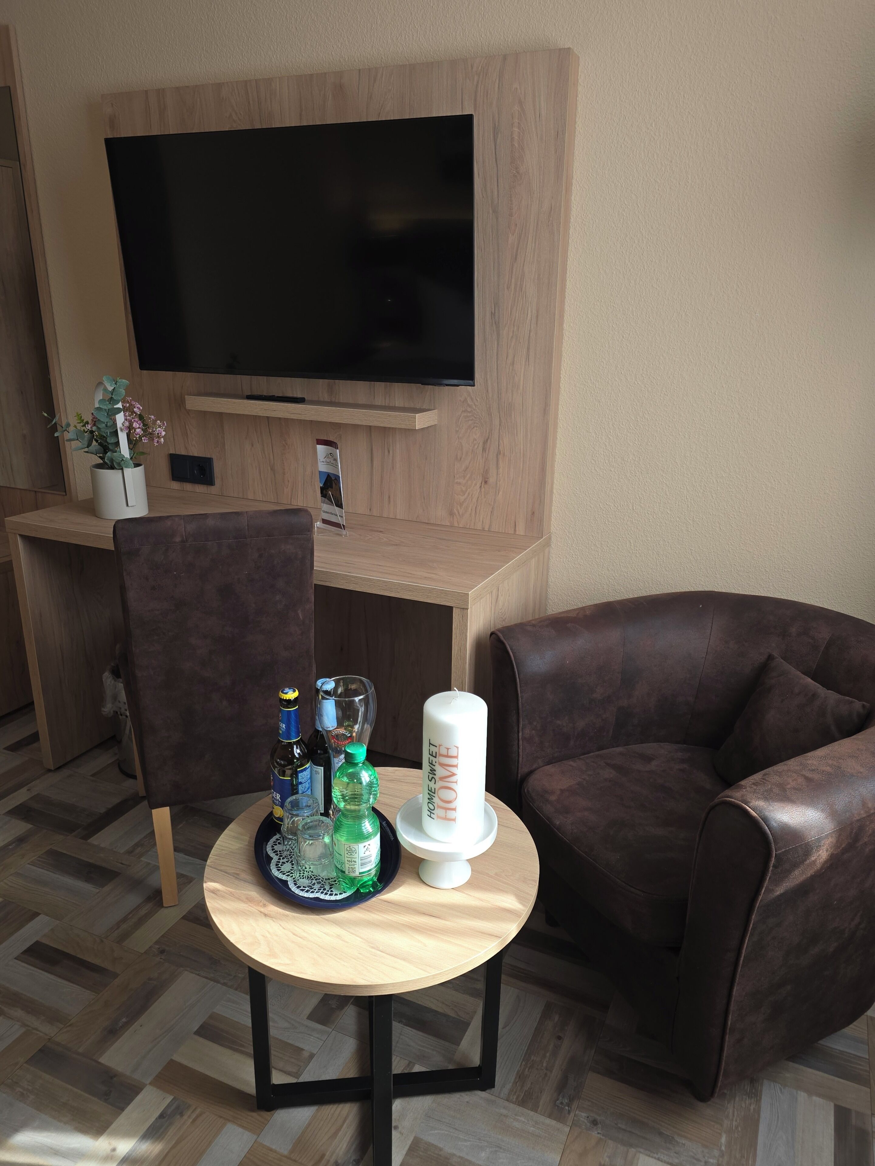 A living room with a flat-screen TV mounted on a wooden wall, a brown leather sofa, a round wooden coffee table with bottles and a candle, and a small potted plant.