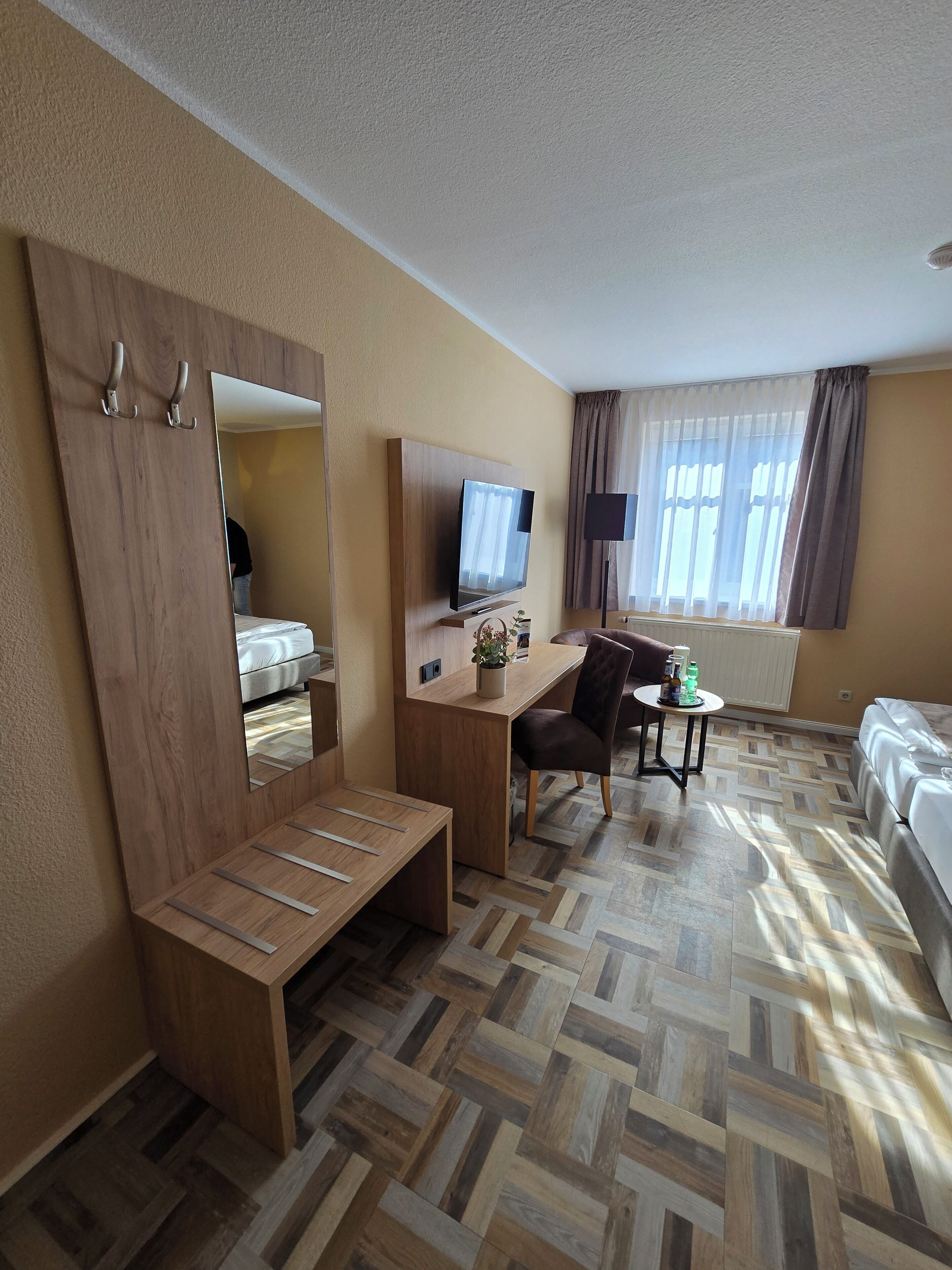 A hotel room with a wooden desk, a chair, a bed, a TV, and a large mirror.