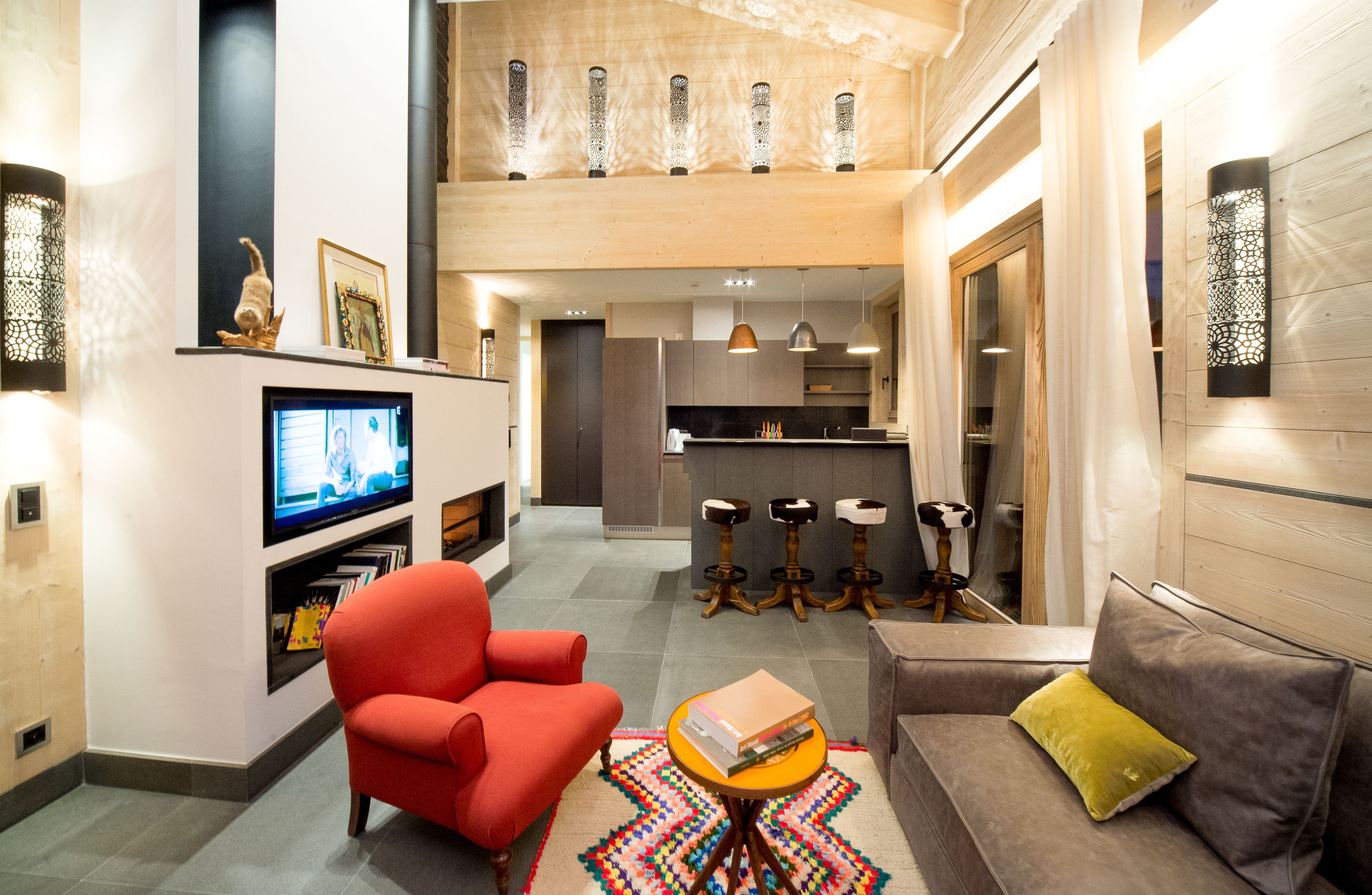 A modern living room with a red armchair, a gray sofa, and a television. There is a kitchen area in the background with a bar and stools.