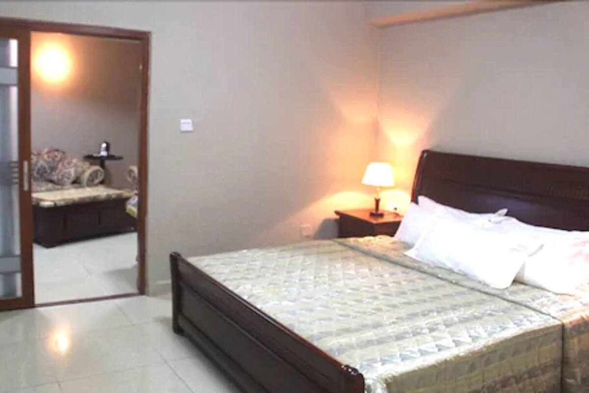 A bedroom with a bed, a nightstand with a lamp, and a bicycle in the background.
