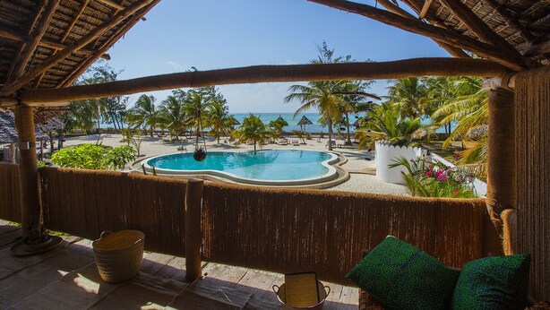 A pool area with a view of the beach and ocean, surrounded by palm trees and a thatched roof structure. - Ujamaa Beach Resort Zanzibar (Makunduchi)