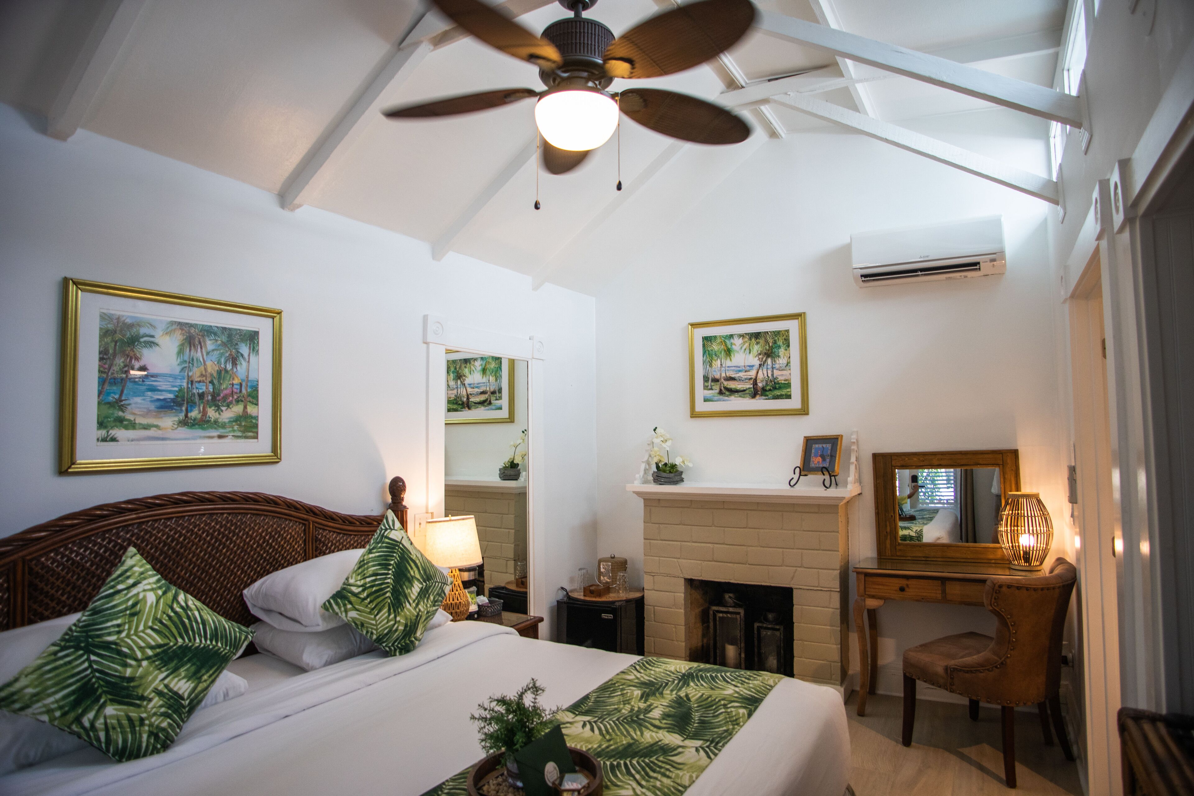 Un dormitorio con cama, escritorio, silla, ventilador de techo, chimenea y dos cuadros en la pared.