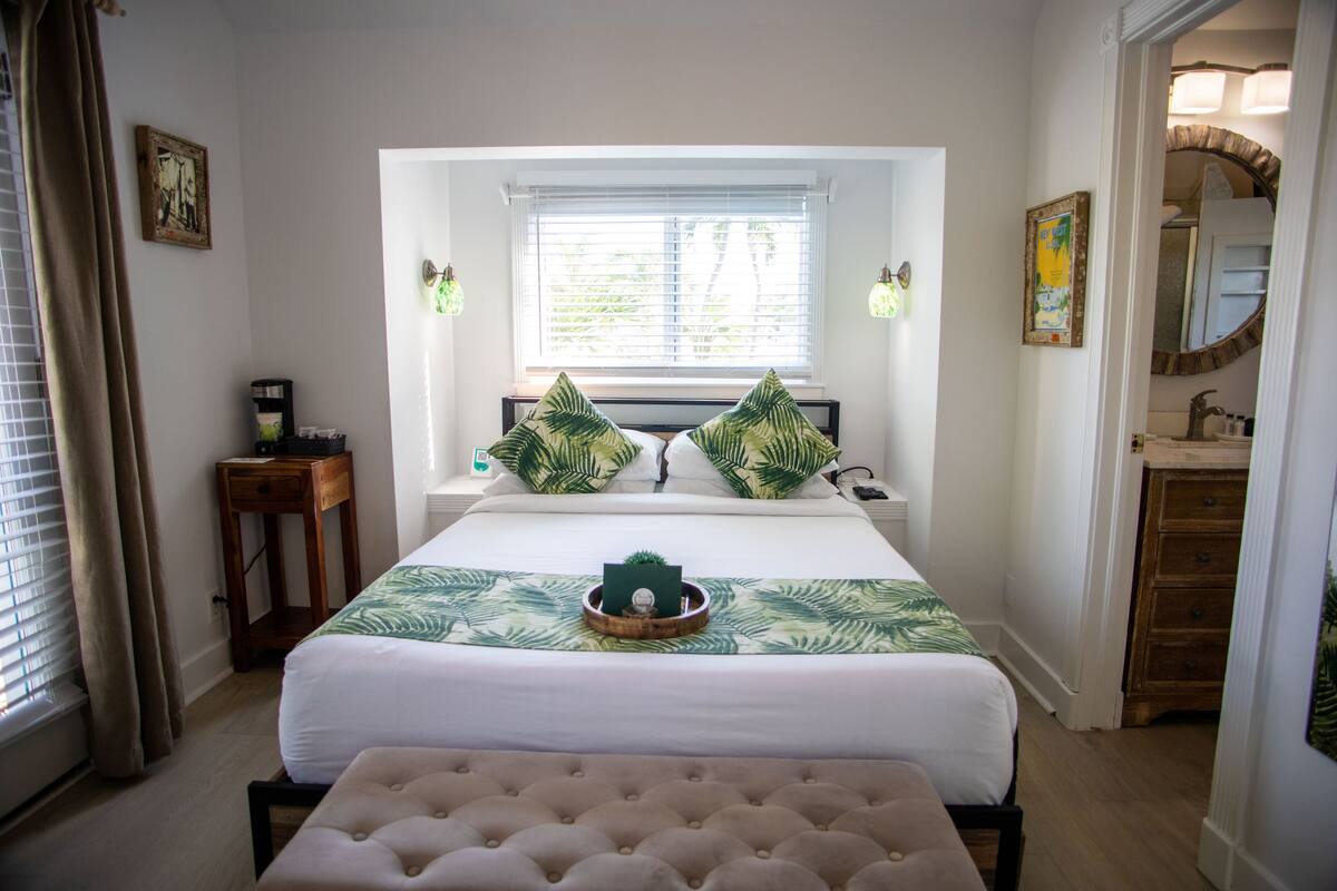 Une chambre à coucher avec un grand lit, une table de chevet, une commode, un banc, une fenêtre avec des stores et un miroir.