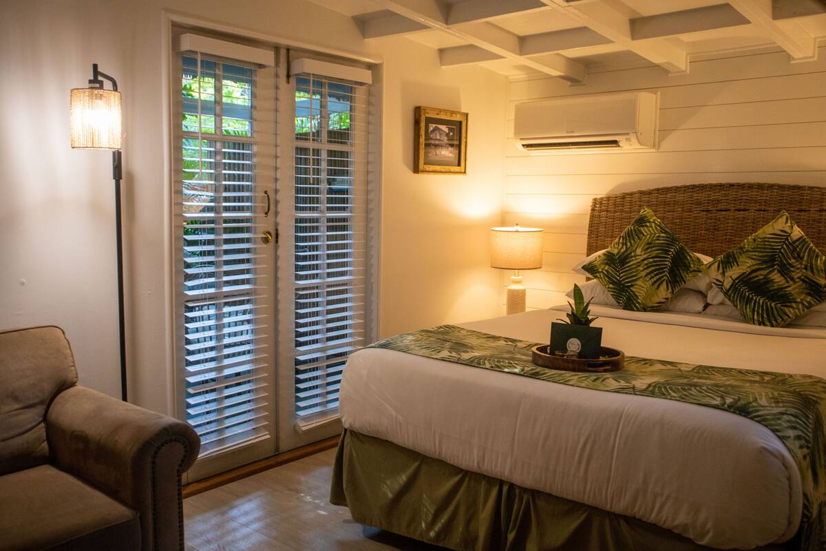 Une chambre à coucher avec un lit, une table de chevet, une lampe, une chaise et une fenêtre avec des stores.