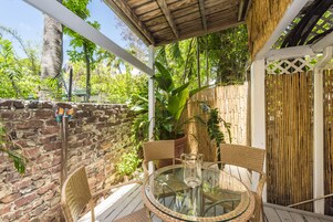 A glass-top table with wicker chairs, a potted plant, and a bamboo fence in a covered outdoor area.