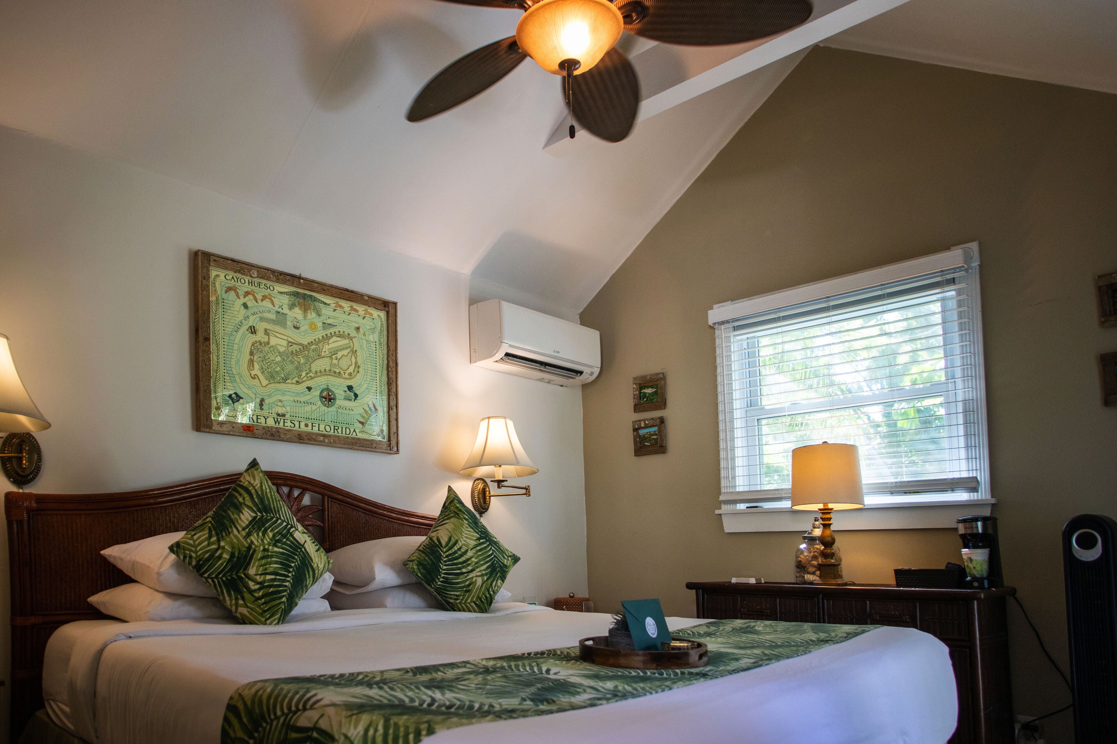 A hotel room with a bed, a ceiling fan, a window with blinds, and wall-mounted air conditioning.