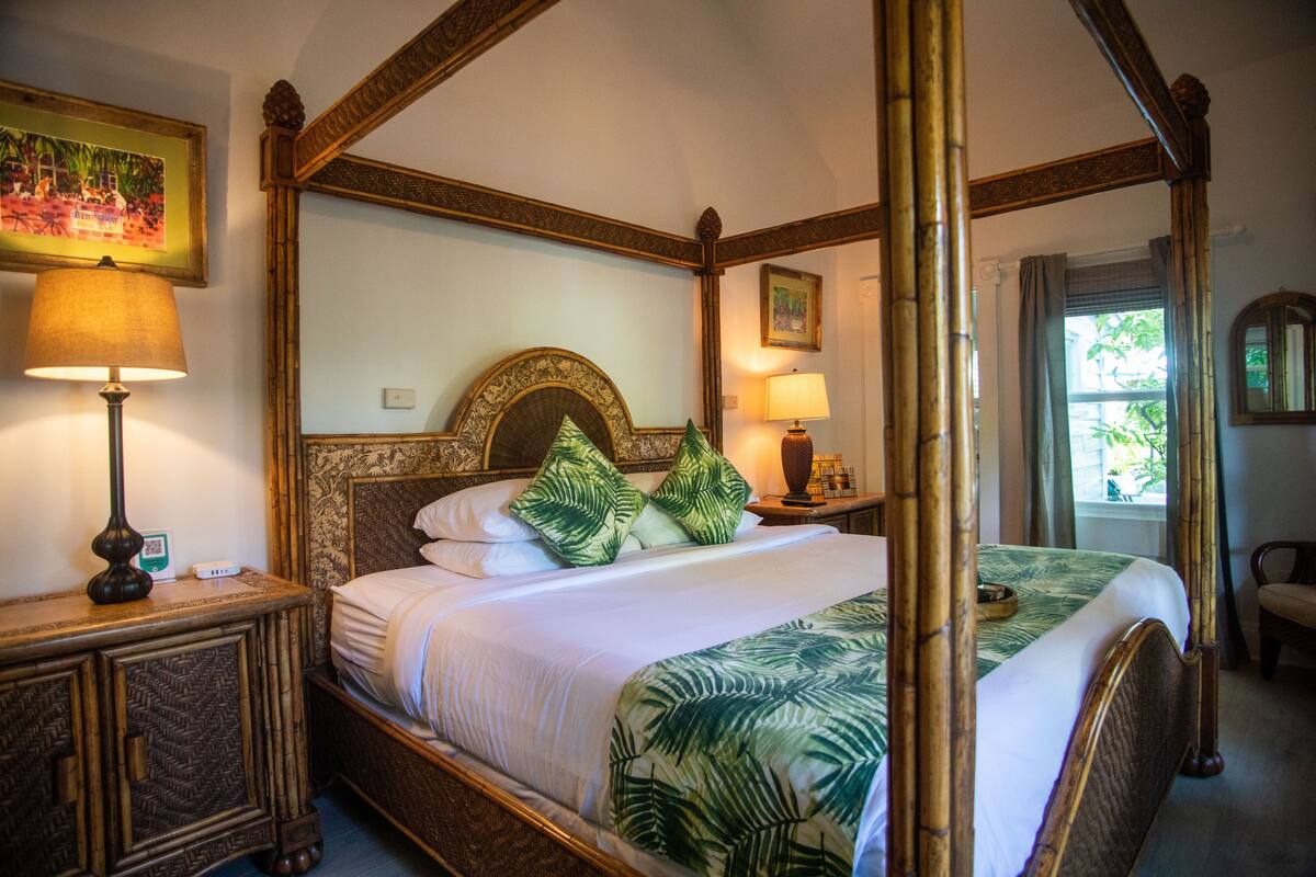 Un lit à baldaquin avec une structure en bois, du linge de lit blanc et une couvre-lit à motifs de feuilles vertes. Il y a deux tables de chevet avec des lampes et une petite horloge sur le côté gauche.
