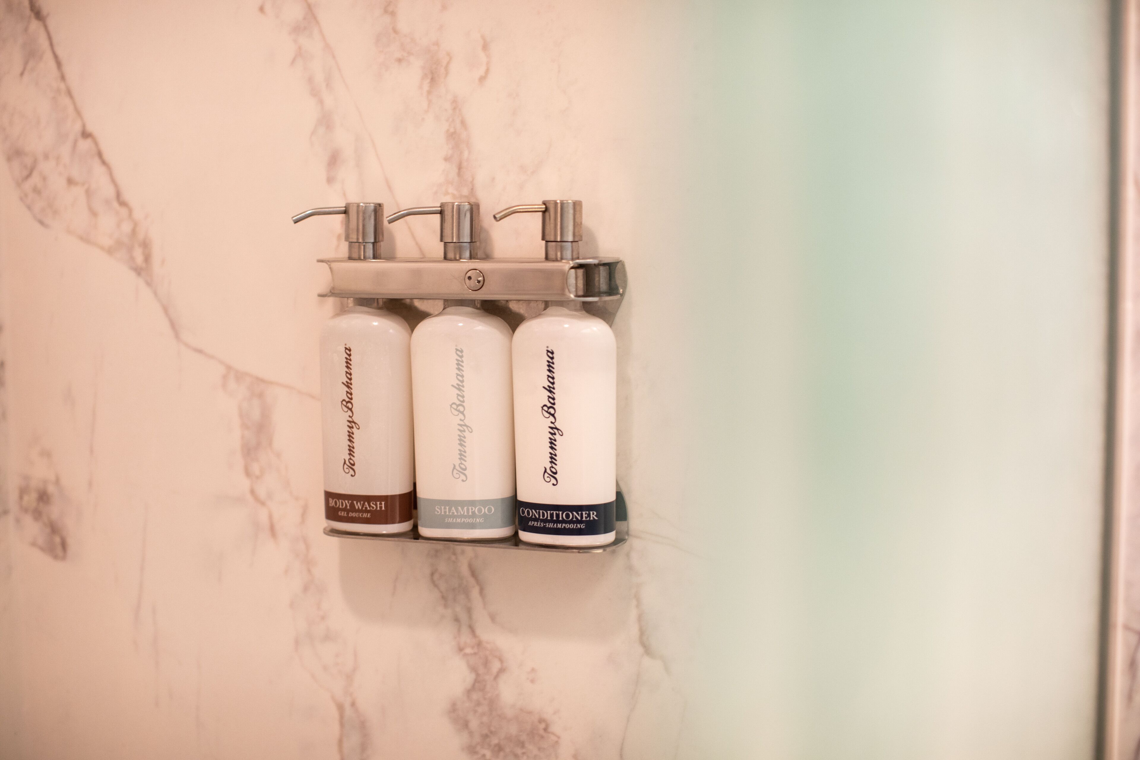 A bathroom with a marble wall and a wall-mounted dispenser holding three bottles of Tommy Bahama body wash, shampoo, and conditioner.