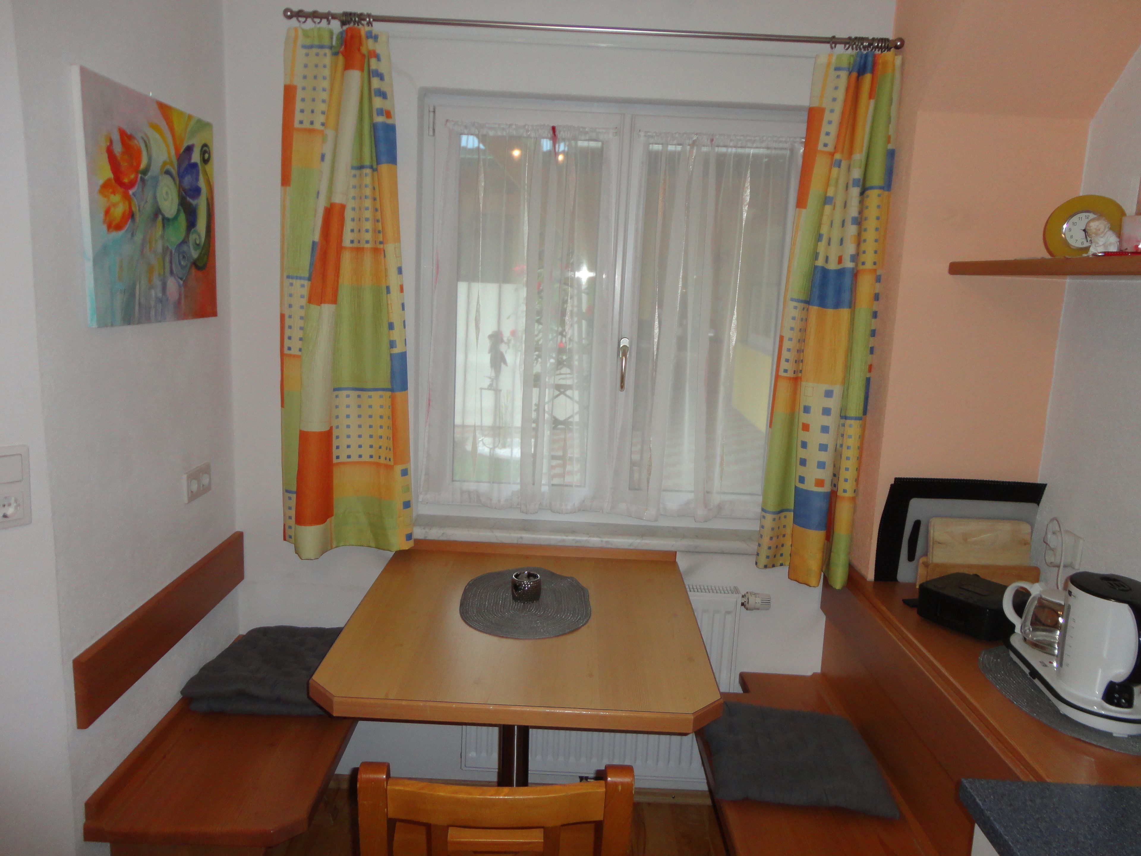 A cozy room with a wooden table, benches, a window with curtains, a painting on the wall, and a shelf with a clock and a cup.
