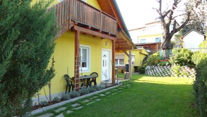 A yellow building with a wooden balcony, a small garden with a lawn and shrubs, and a wooden fence. - Ferienwohnungen Fam. Pabst (Maria Lankowitz)