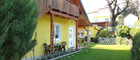 A yellow building with a wooden balcony, a small garden with a lawn and shrubs, and a wooden fence.