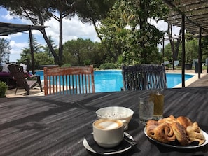 A table with a cup of coffee, a glass of water, and pastries, overlooking a pool with chairs and umbrellas. - Almanuda Naturist Retreat (Sutri)