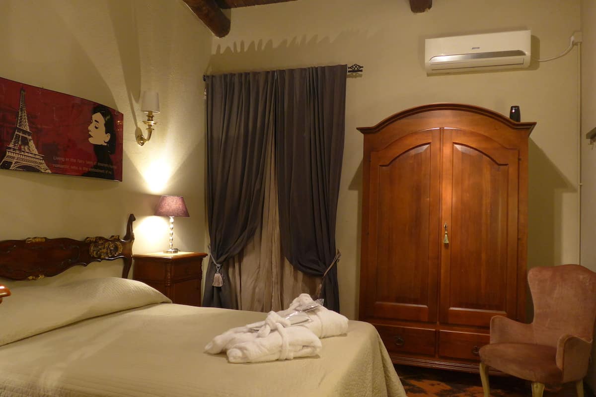 A hotel room with a bed, a wooden wardrobe, a chair, a nightstand with a lamp, and a wall-mounted air conditioner.