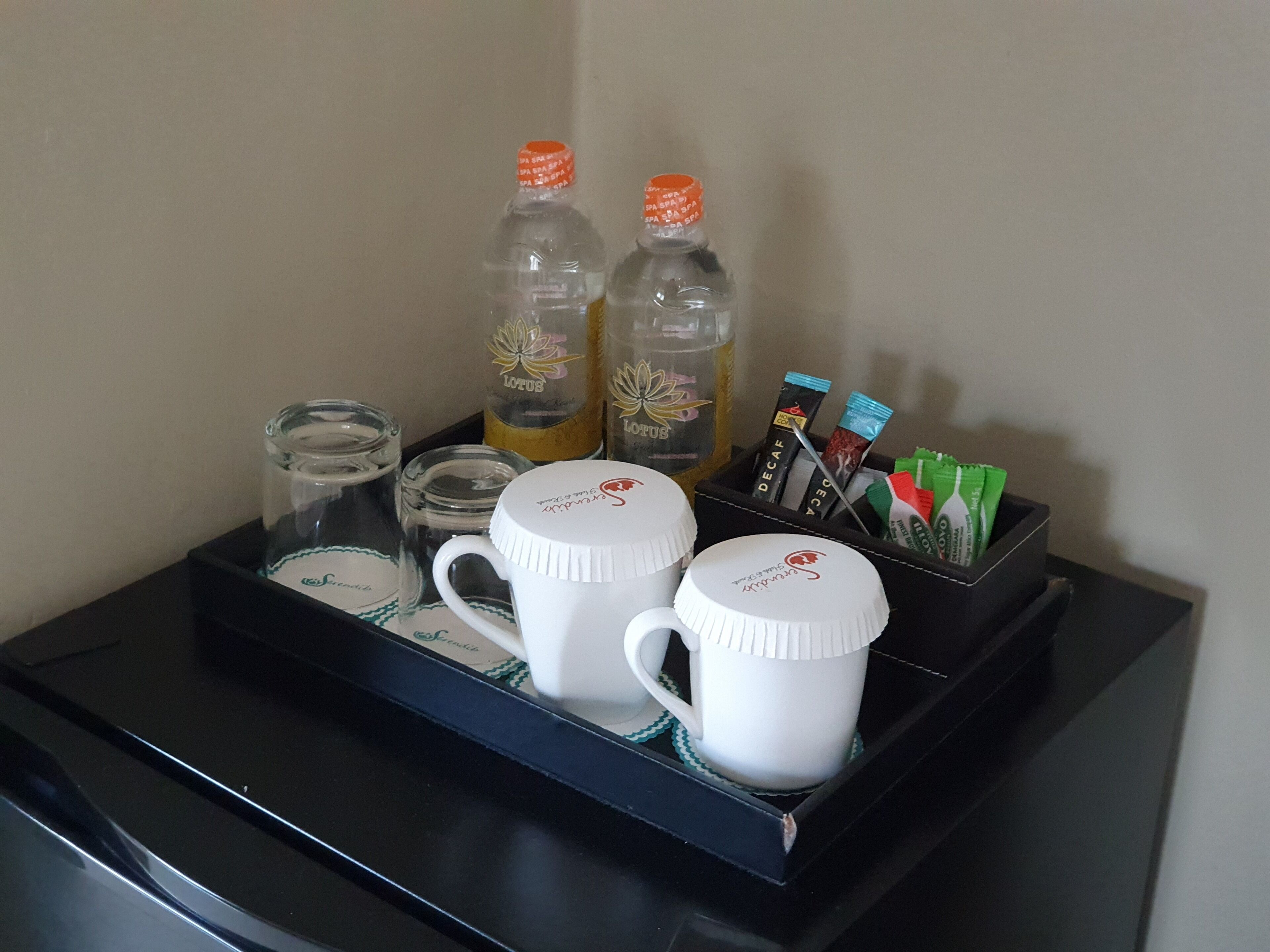 Un plateau contenant deux tasses blanches, deux bouteilles en verre et un récipient renfermant divers objets, notamment un stylo et un petit paquet.