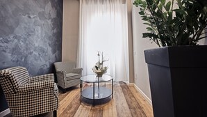Una sala moderna con una mesa de centro redonda de vidrio, dos sillones estampados, una planta grande en maceta y vistas de una cortina blanca y una ventana.