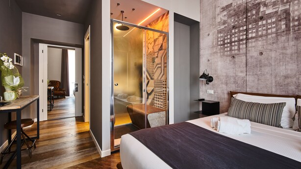 Habitación de hotel moderna con una cama grande, cabina de ducha de vidrio y un área de comedor visible a través de una puerta abierta.