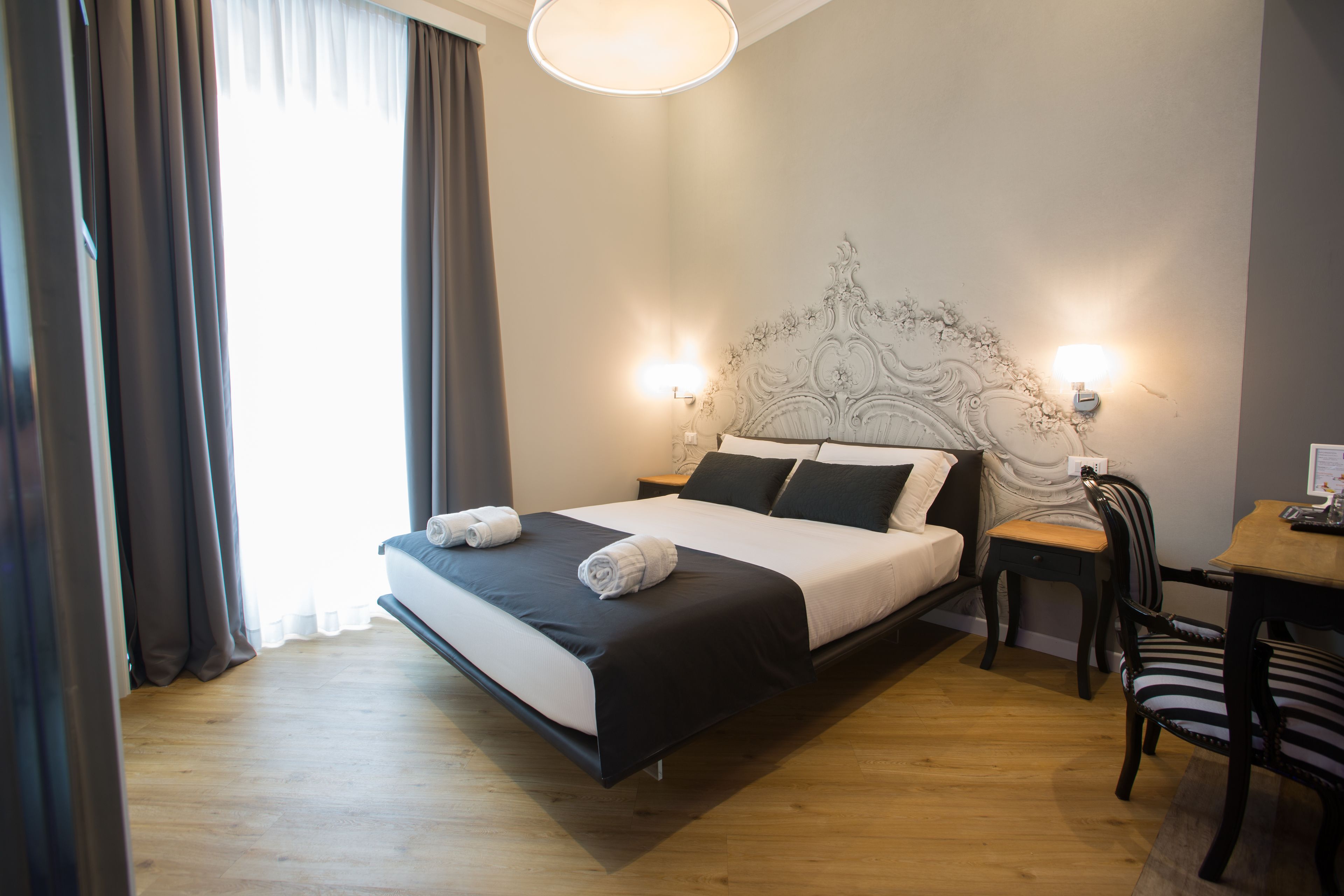 Una cama bien hecha con edredón blanco y negro, dos lámparas de noche, una mesita de noche de madera, una silla a rayas y una pequeña mesa con una lámpara.