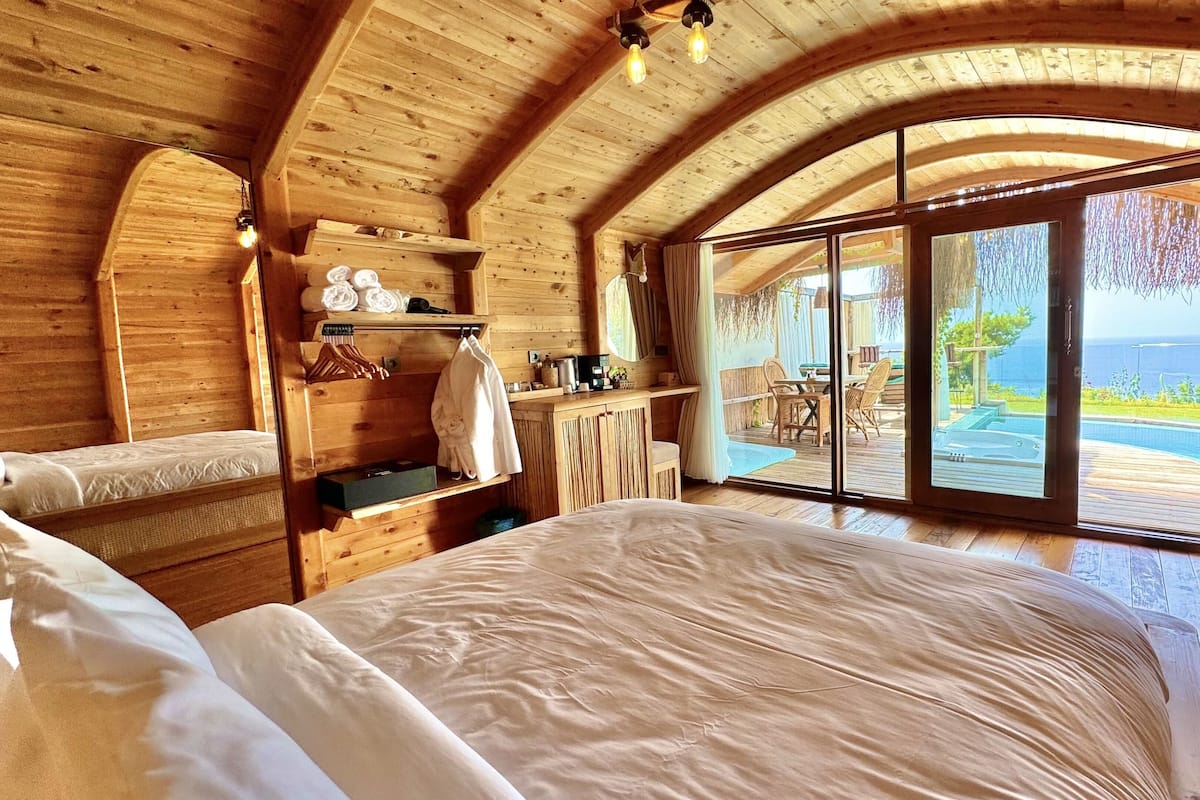 A wooden cabin bedroom with a large bed, a small table, and a view of the ocean through a sliding glass door.