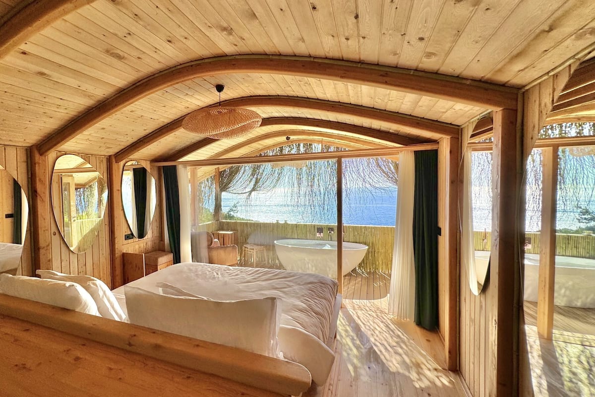 A wooden cabin with a large bed, a small table, and a view of the outdoors.