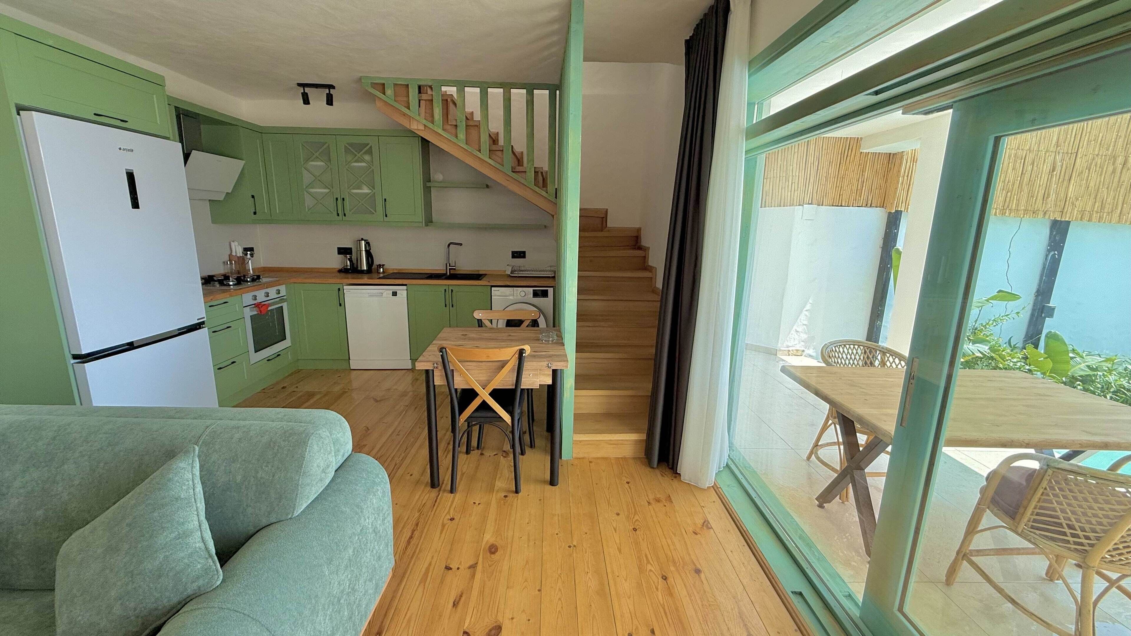 A compact kitchen with green cabinets, a dining area with a wooden table and chairs, and a staircase leading to an outdoor area.