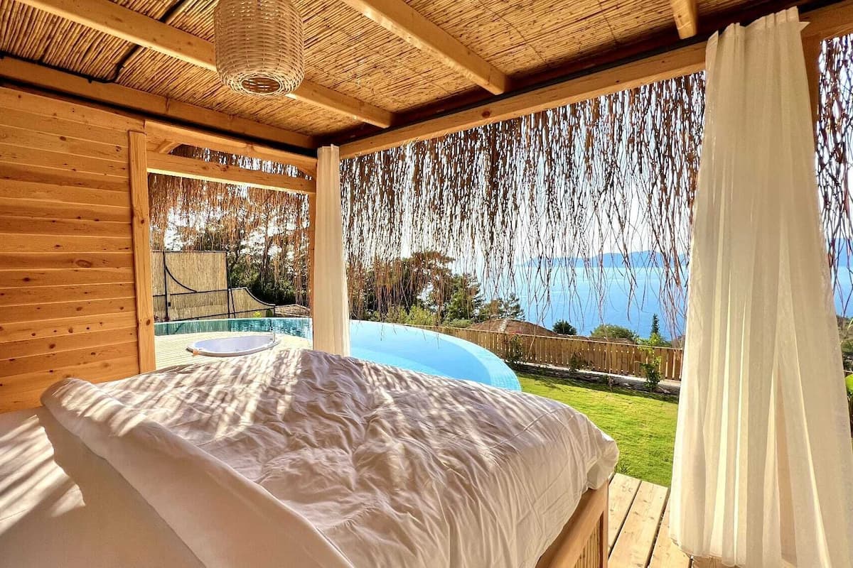 A wooden room with a bed, a hanging light fixture, and a view of a pool and greenery.