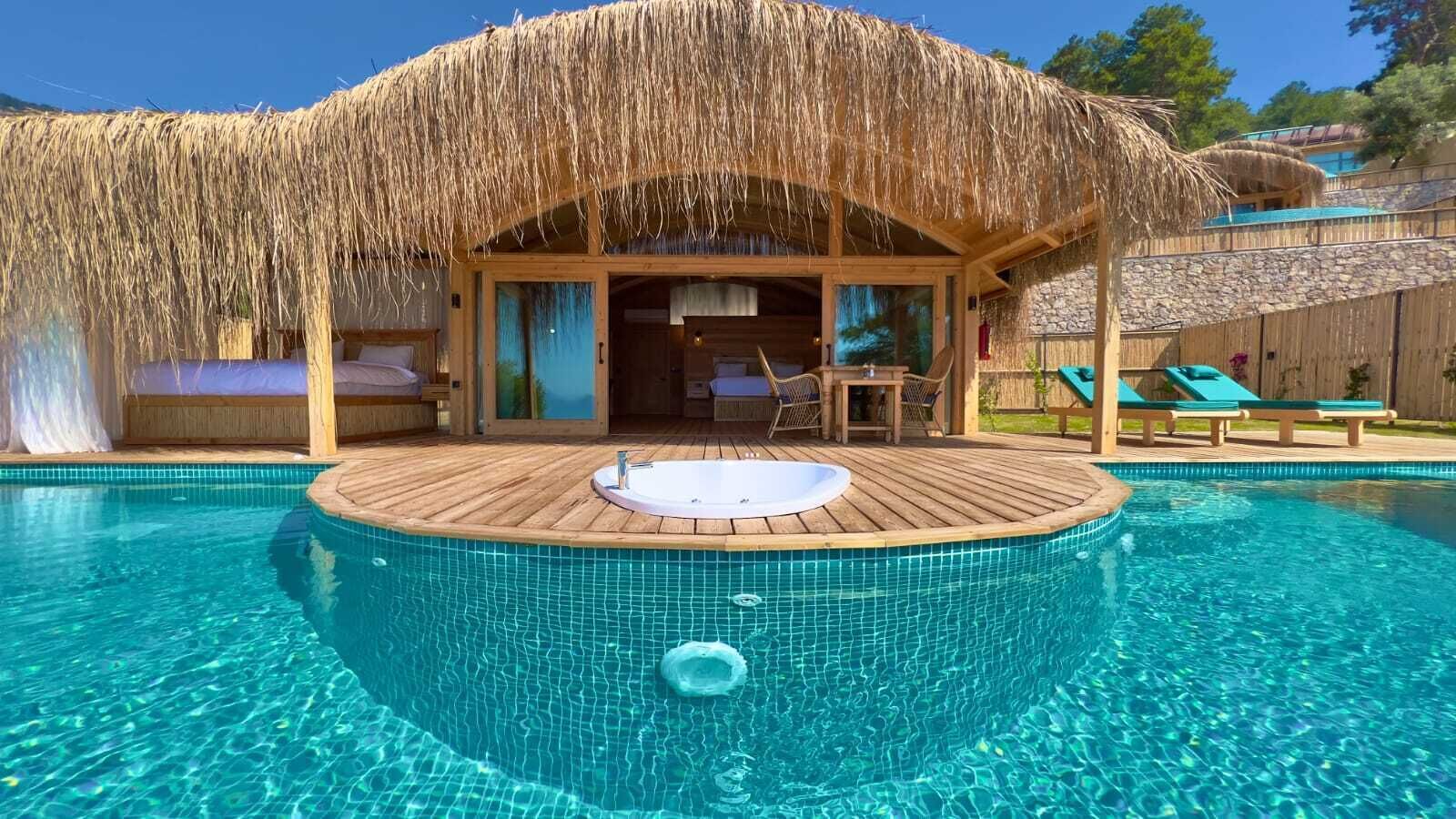 Een zwembad met een jacuzzi en ligbedden, omgeven door een houten terras en een constructie met rieten dak.