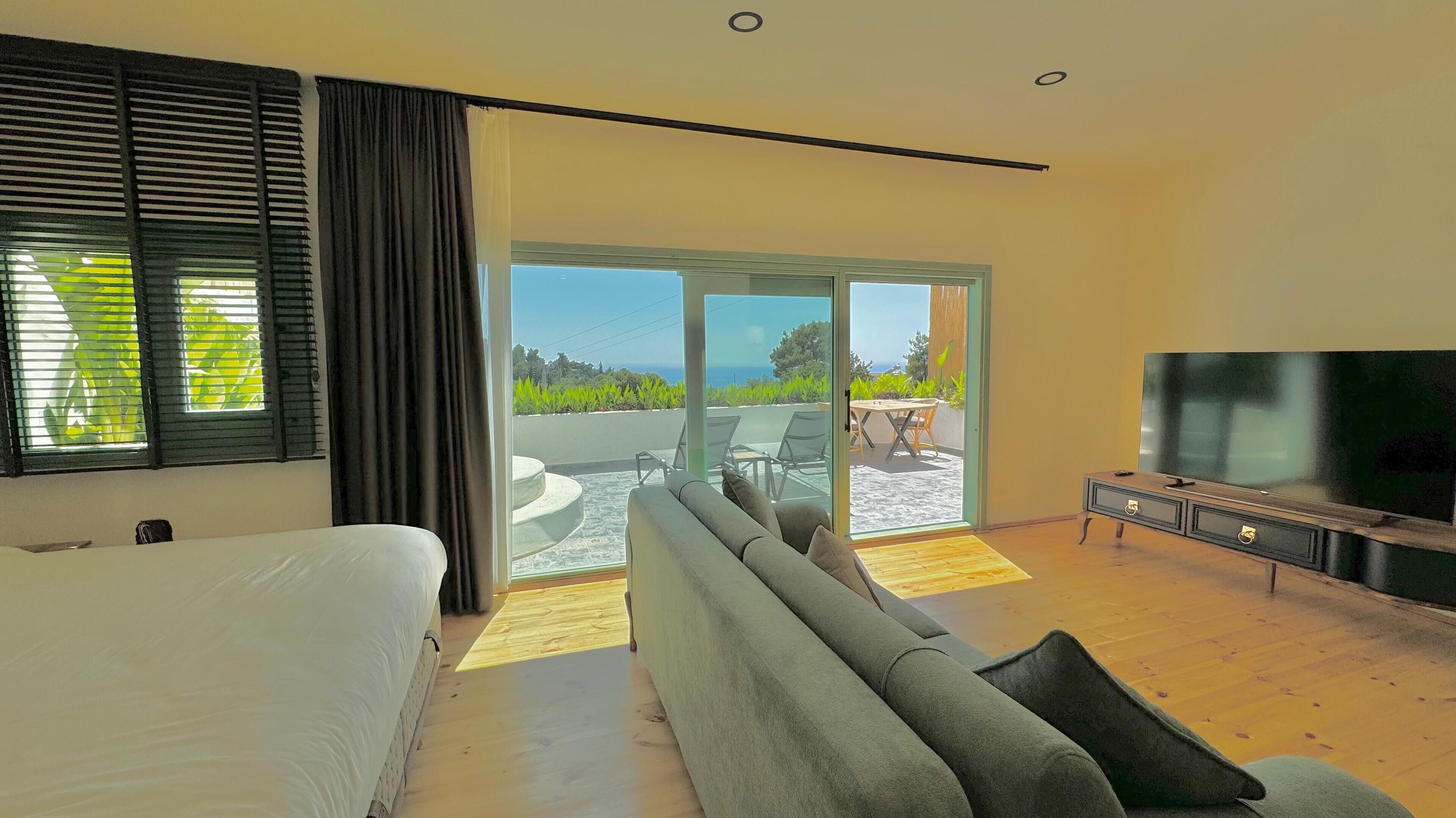 Un dormitorio moderno con una cama grande, un sofá, un televisor y un ventanal corredera que da a un balcón con mobiliario exterior.