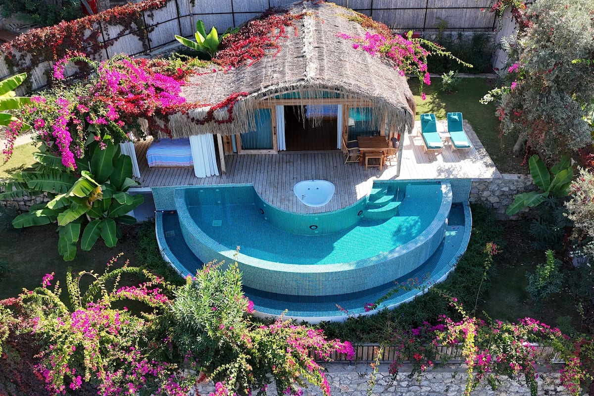 A circular infinity pool with a wooden deck and lounge chairs, surrounded by lush greenery and flowering plants.