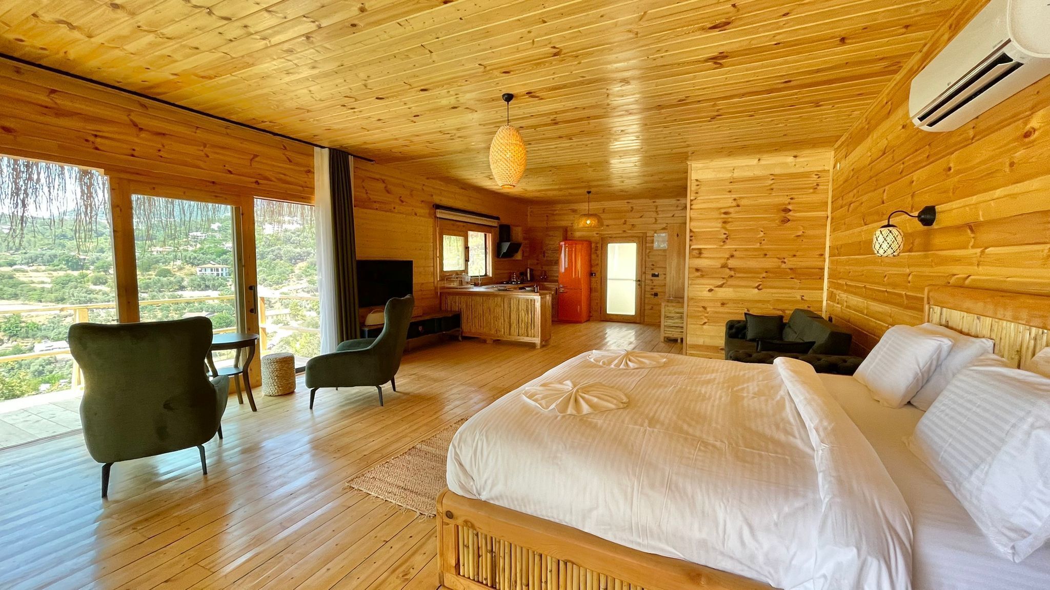 A wooden cabin bedroom with a large bed, a TV, a kitchen area, and a view of the outdoors.