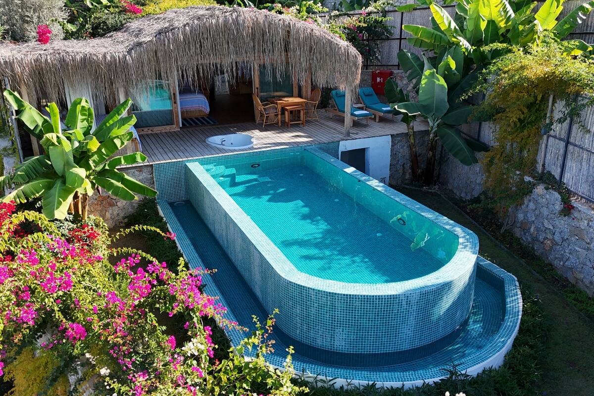 A swimming pool with a tiled deck, a thatched-roof structure, and lush greenery.