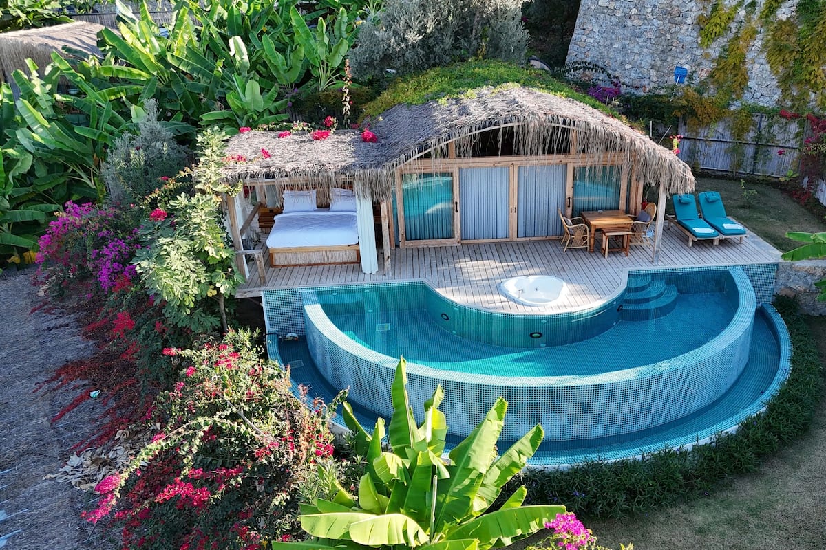 A thatched-roof hut with a bed and a small table, surrounded by a swimming pool and lush greenery.