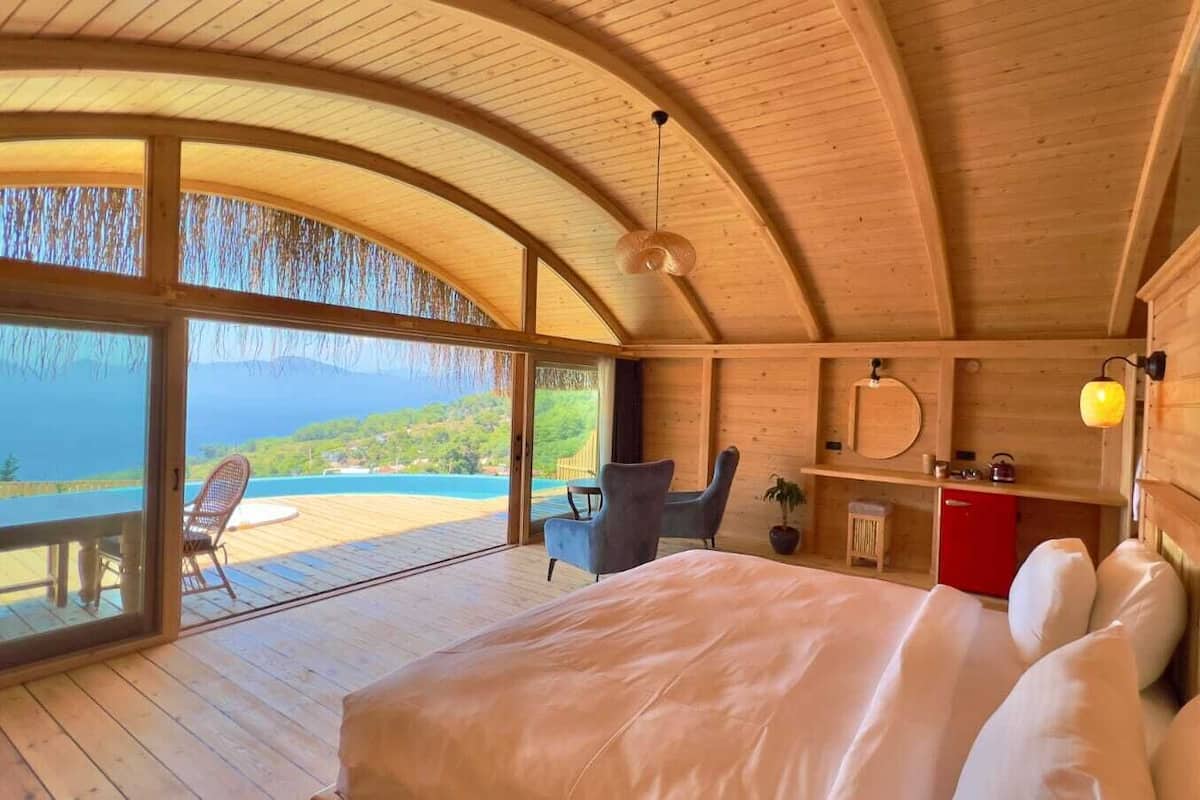 A wooden cabin with a large bed, a desk, and a chair. There is a view of the ocean and mountains through the windows.