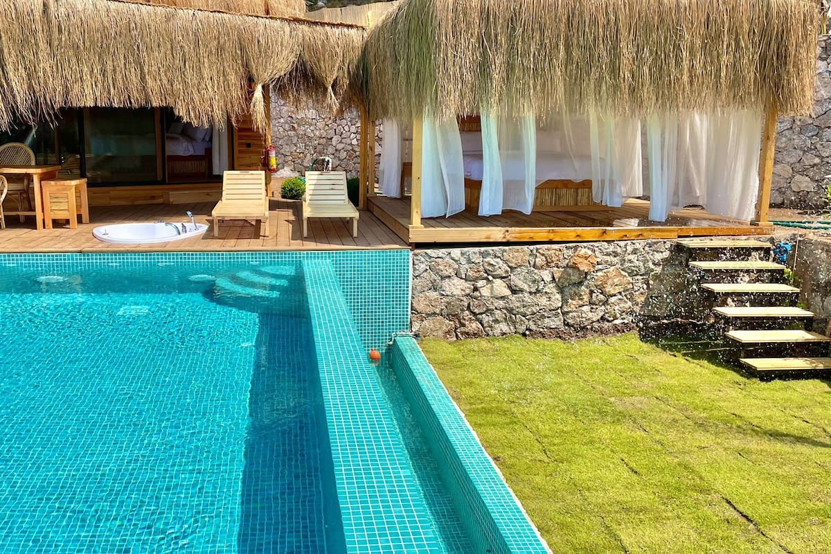 A swimming pool with a stone wall and a thatched roof area with seating.