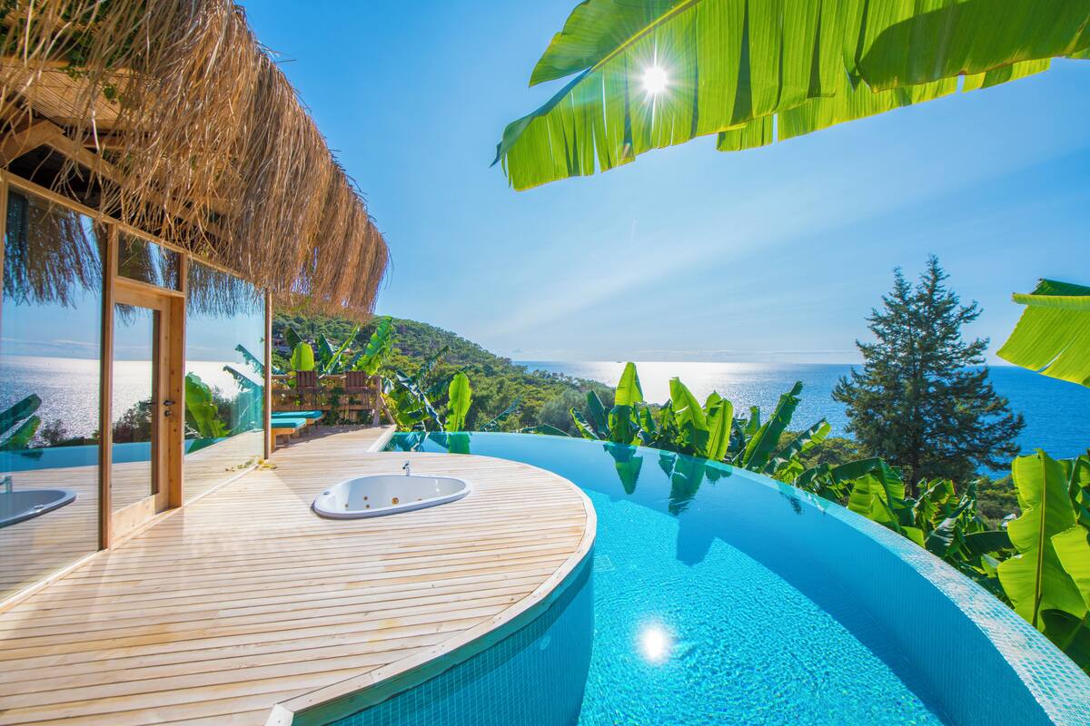 A infinity pool with wooden deck and a hot tub, surrounded by tropical plants and overlooking a scenic view.