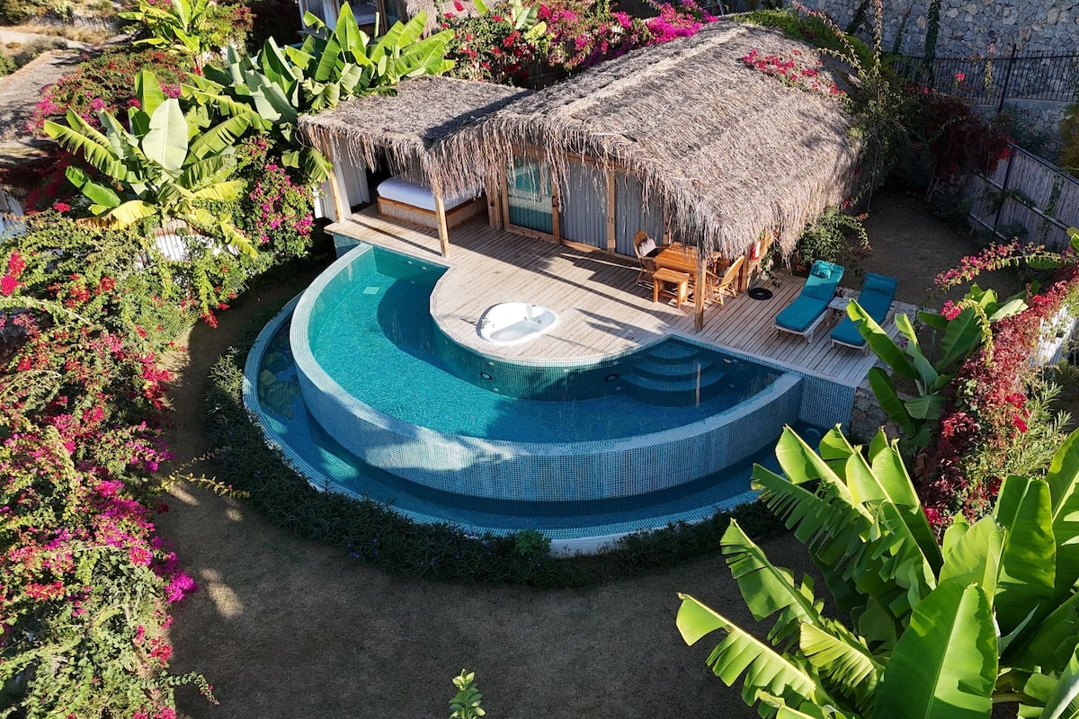 A circular infinity pool with a hot tub, surrounded by lush greenery and a thatched-roof structure.