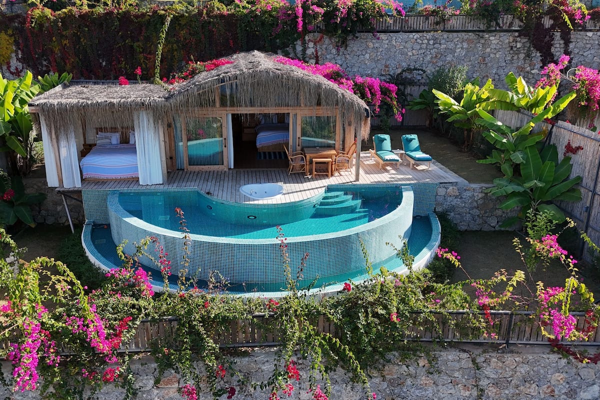 A thatched-roof bungalow with a private pool, surrounded by lush vegetation and a stone wall.