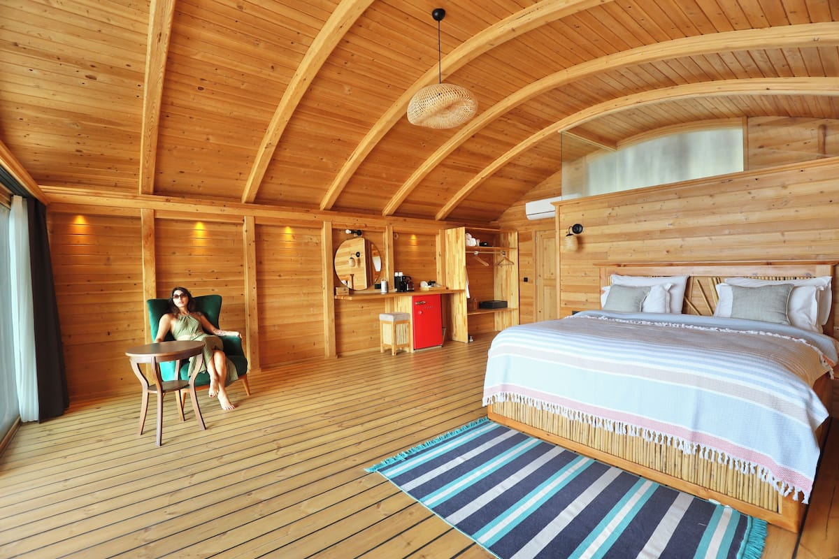 A wooden cabin interior with a bed, a chair, a small table, and a kitchenette.