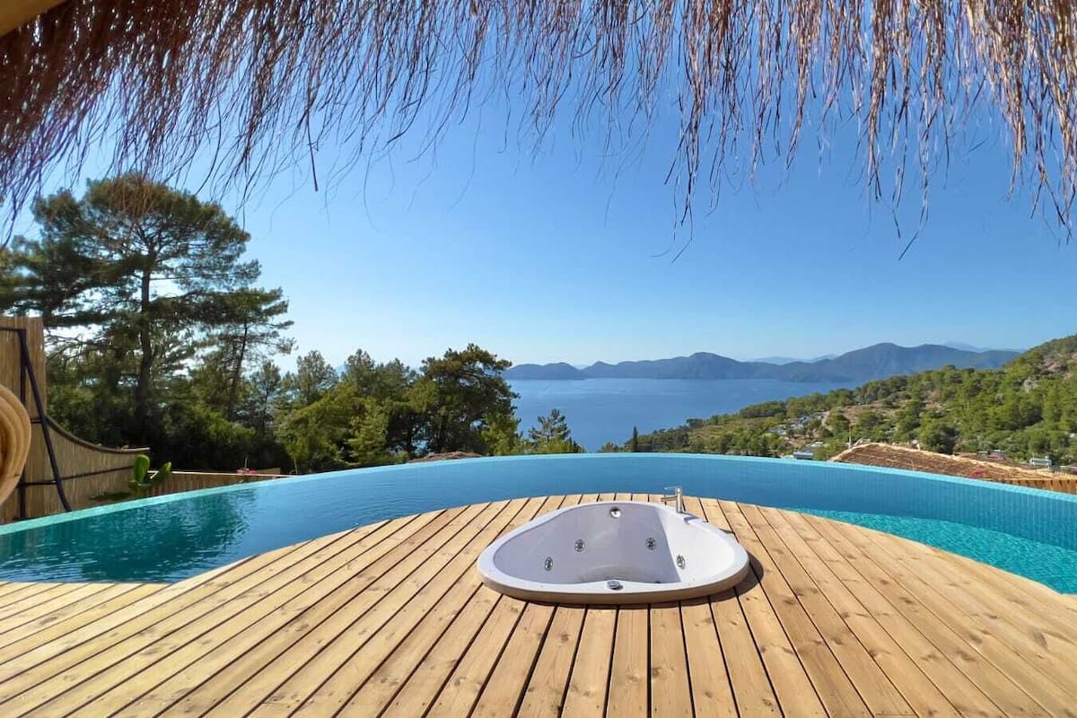 An infinity pool with a hot tub, surrounded by wooden decking, overlooking a scenic view of the sea and hills.