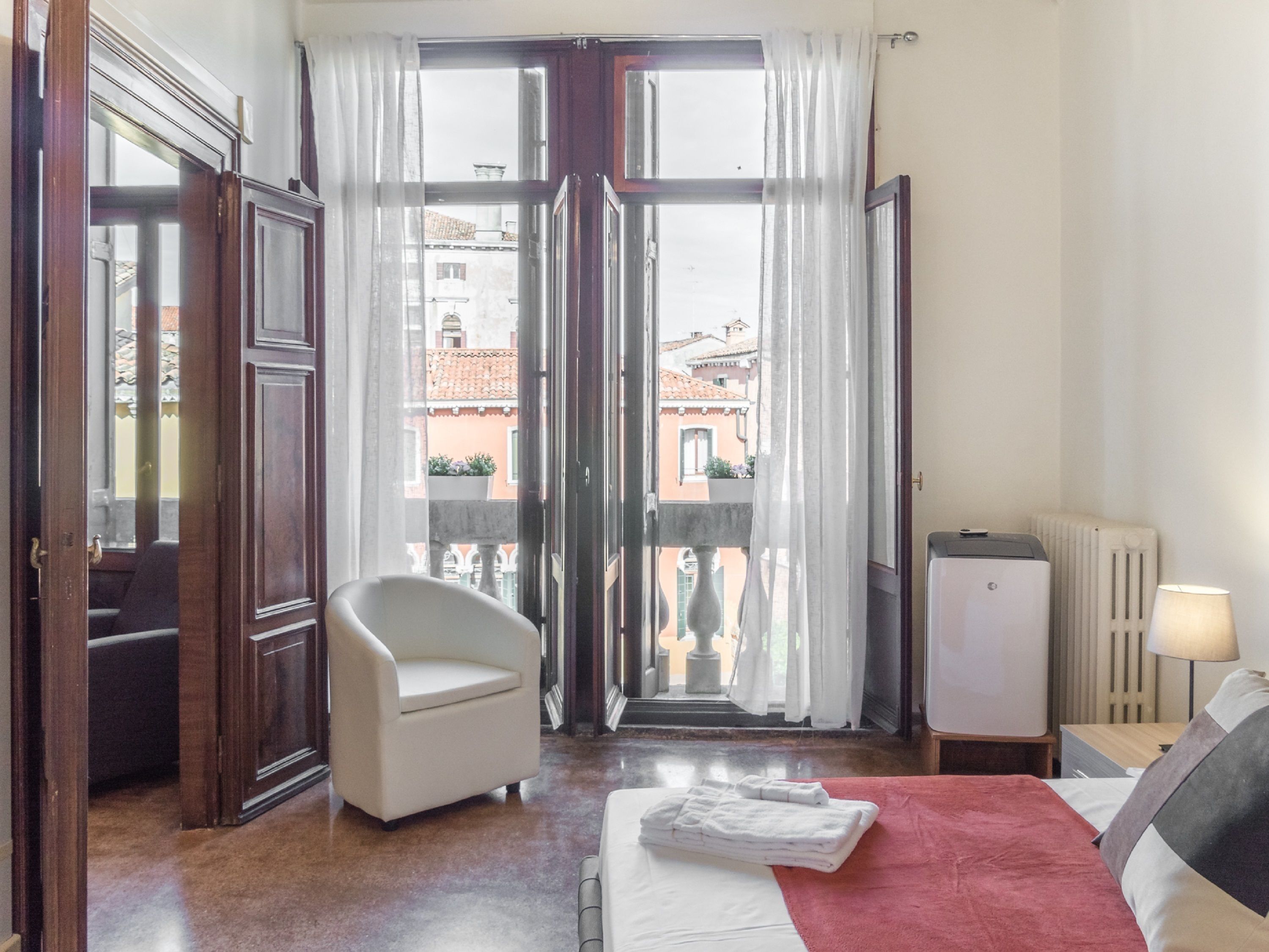 Ein Hotelzimmer mit Bett, Stuhl, Heizkörper und Blick auf Gebäude durch die geöffnete Tür.