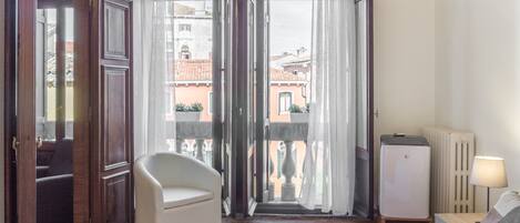 A hotel room with a bed, a chair, a radiator, and a view of buildings through the open door.