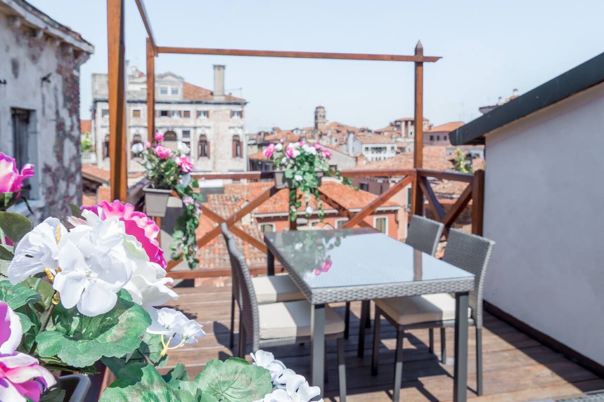 A rooftop terrace with a glass table and chairs, surrounded by potted flowers.