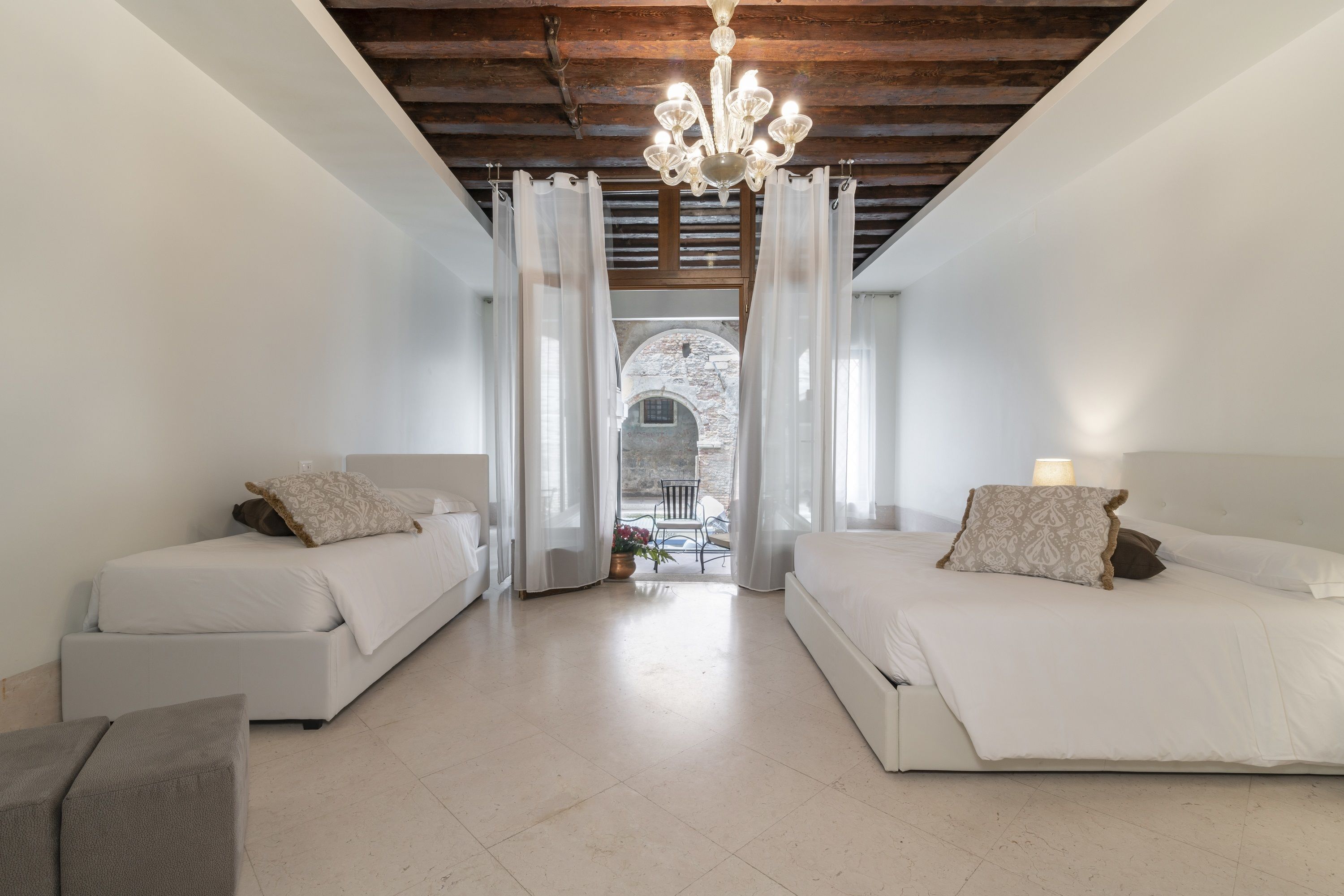 A bedroom with two beds, a chandelier, and a view of an outdoor patio through a window.