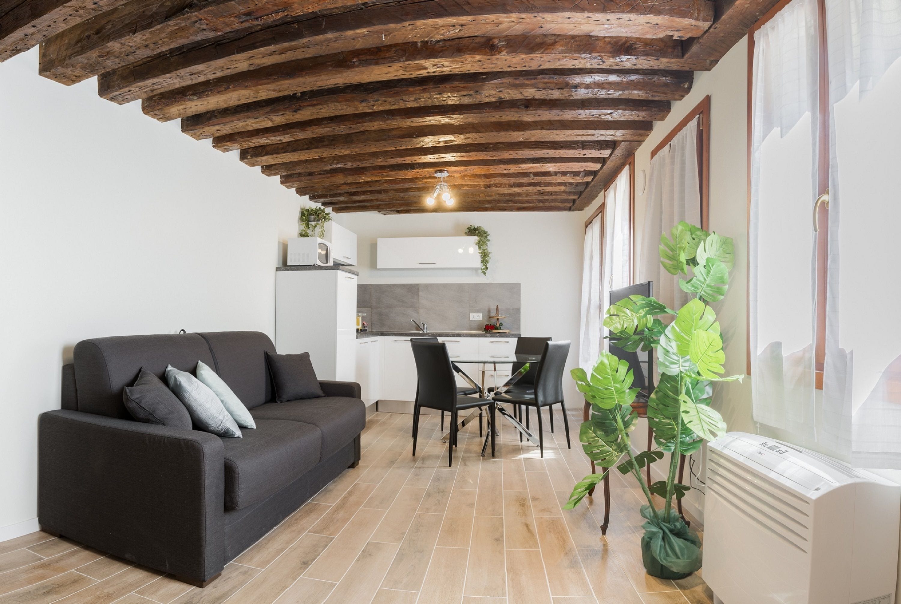 Un salon moderne avec un canapé foncé, une table à manger avec des chaises, une cuisine en arrière-plan et une grande plante.