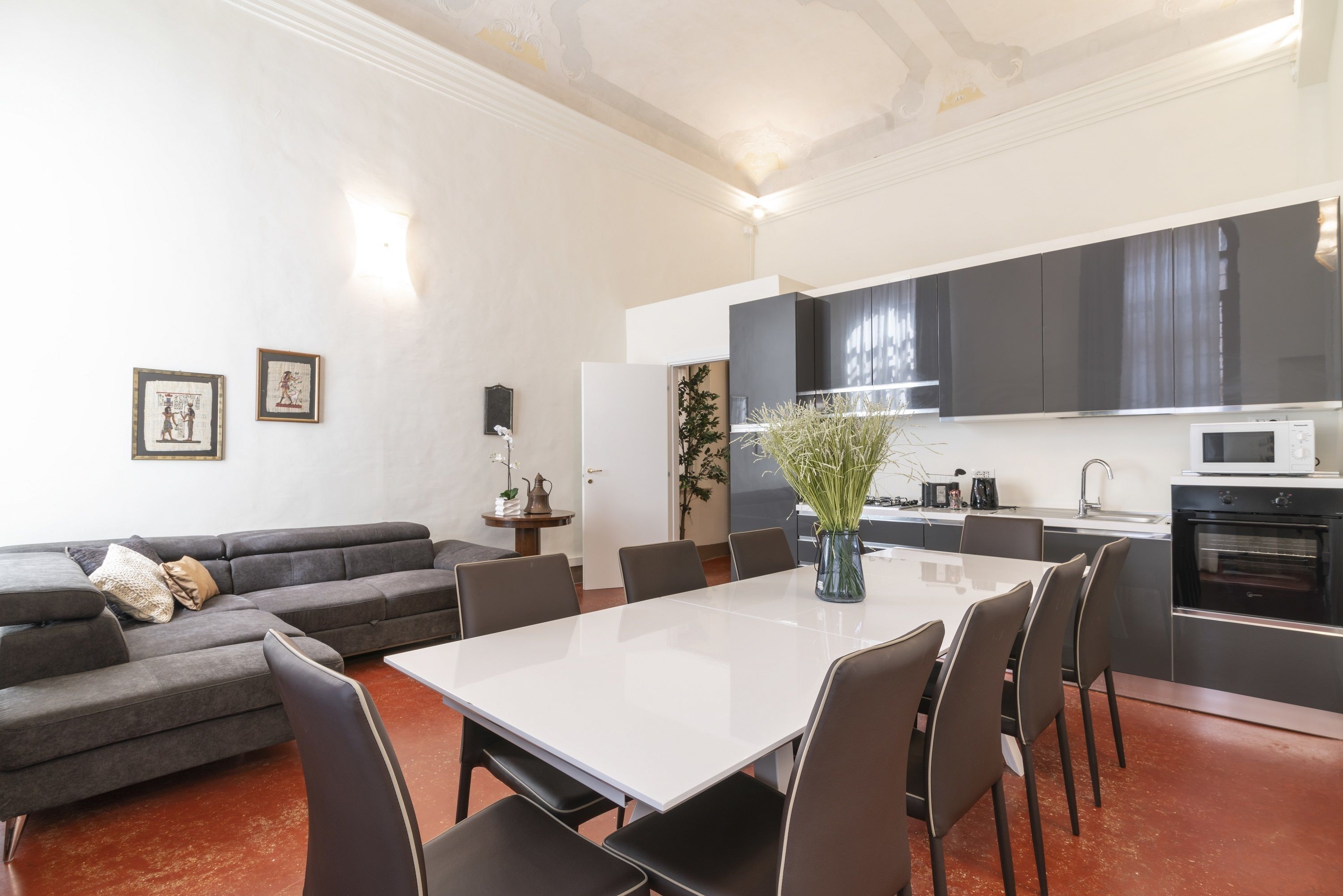 Ein modernes Wohnzimmer mit einem grauen Ecksofa, einem weißen Esstisch und einer Küchenzeile mit dunklen Schränken und einer Mikrowelle.