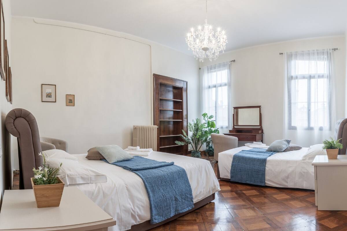 A hotel room with two beds, a wooden headboard, a chandelier, a radiator, a mirror, and a small table with a plant.