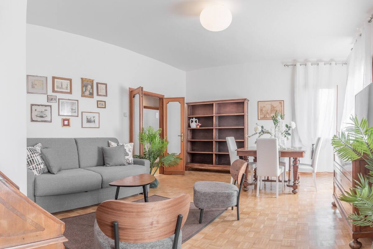 A living room with a grey sofa, wooden coffee table, and chairs. There is a bookshelf, a dining table with chairs, and a potted plant.