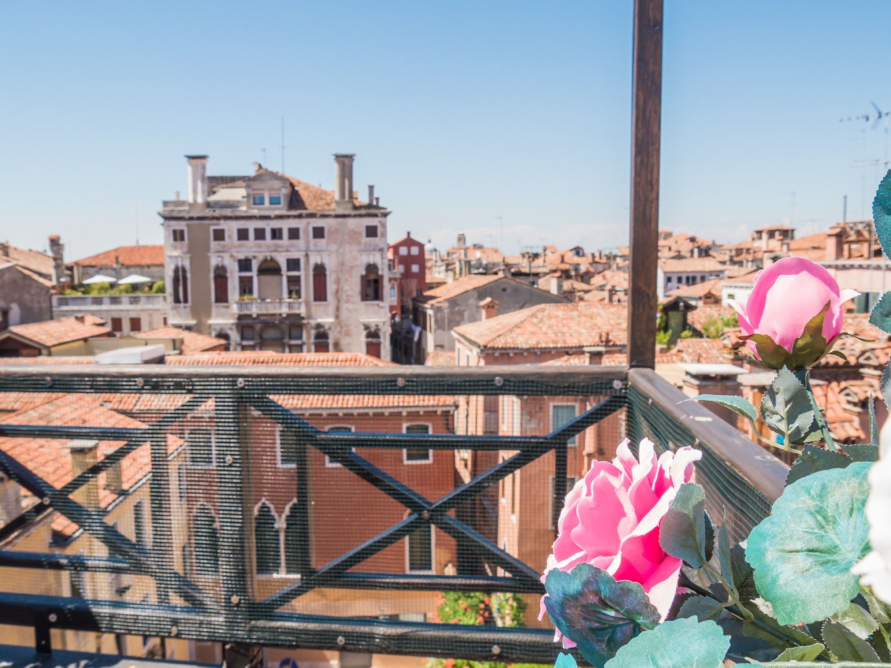 Un balcone con vista su un edificio storico, caratterizzato da un tetto in tegole, una ringhiera in metallo e un fiore rosa in primo piano.