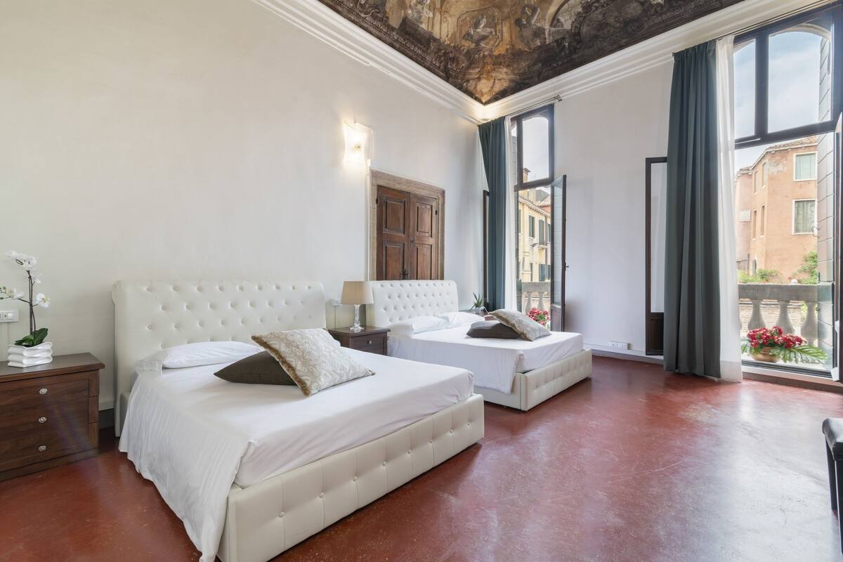 A bedroom with two beds, a wooden nightstand, a wall-mounted lamp, and a large window with curtains.