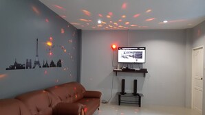 A living room with a brown sofa, a flat-screen TV mounted on the wall, and a ceiling light fixture. - Baan Samart 1 Pool Villa Hua Hin (Hua Hin)