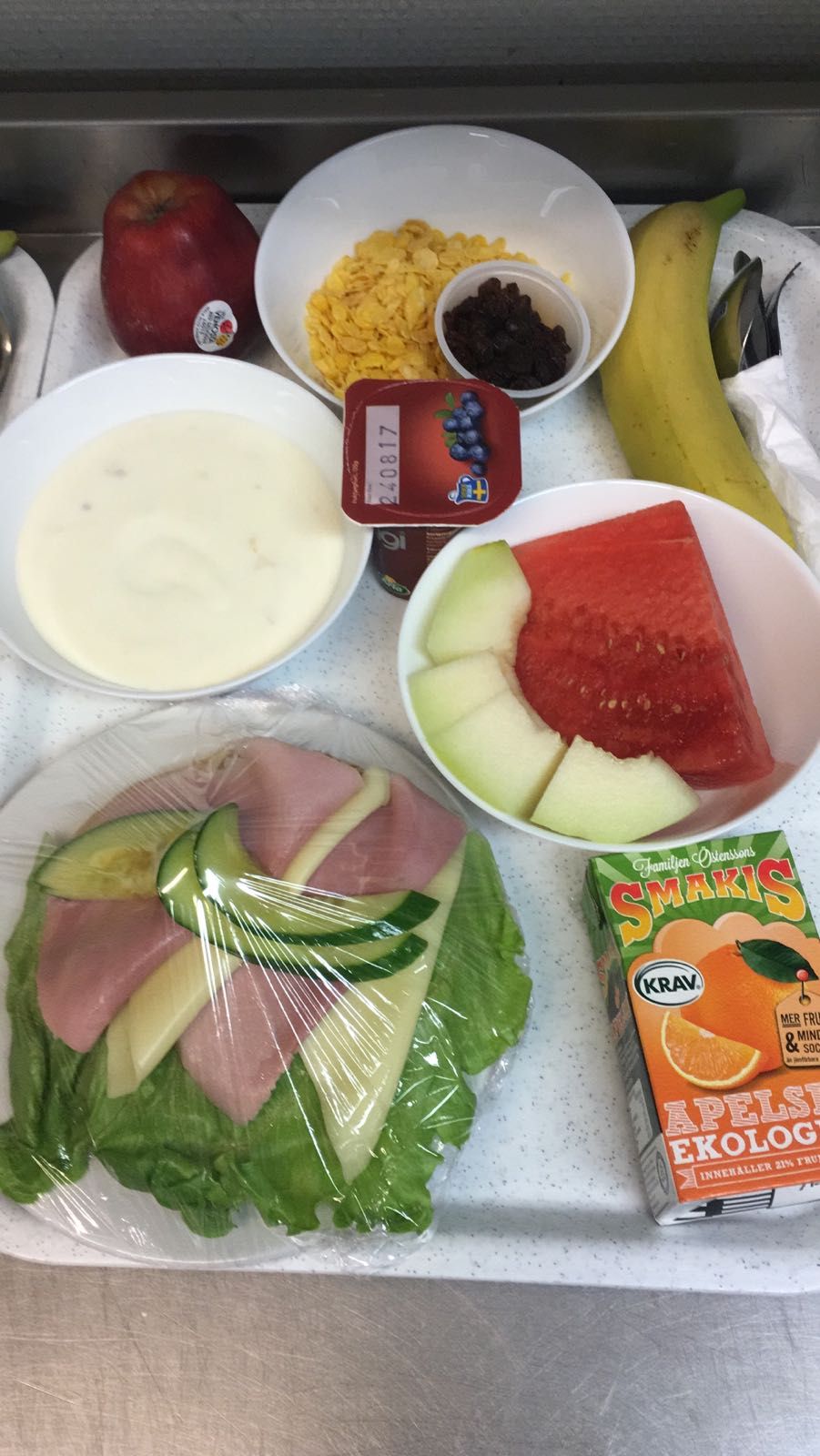 Un refrigerador con varios alimentos, incluyendo una sandía, un plátano, un tazón de cereal, un envase de yogur, un paquete de queso y una caja de jugo de naranja.